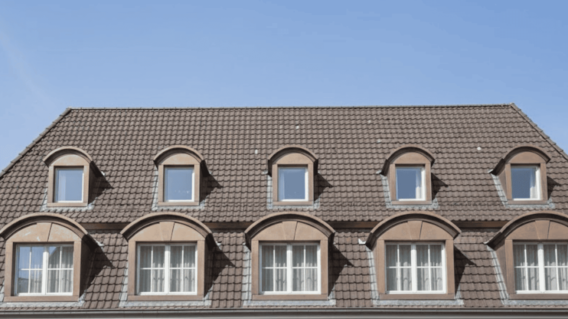 Multiple arched dormer windows aligned across a tiled residential roof under a blue sky