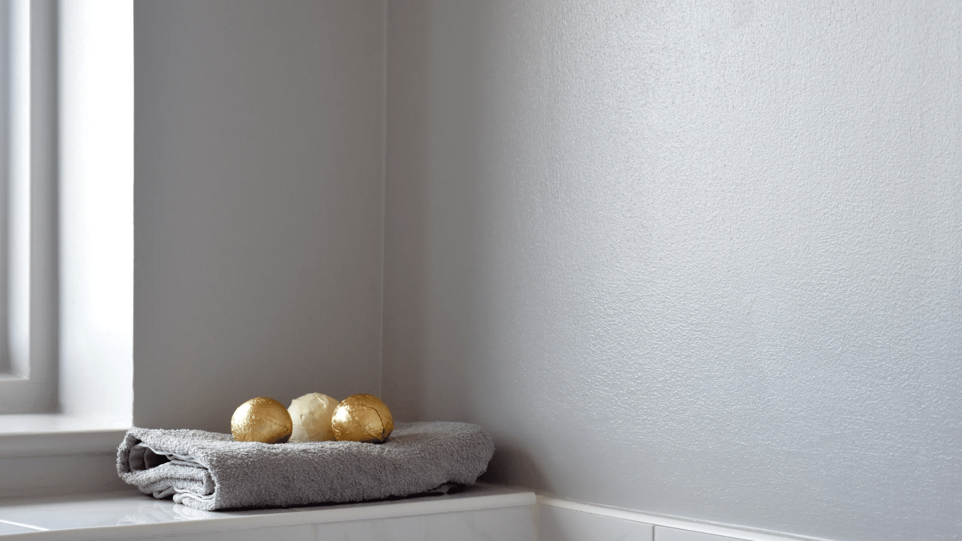 Close up of a gray textured wall in a bathroom with a folded towel and three round gold items on a white marble ledge