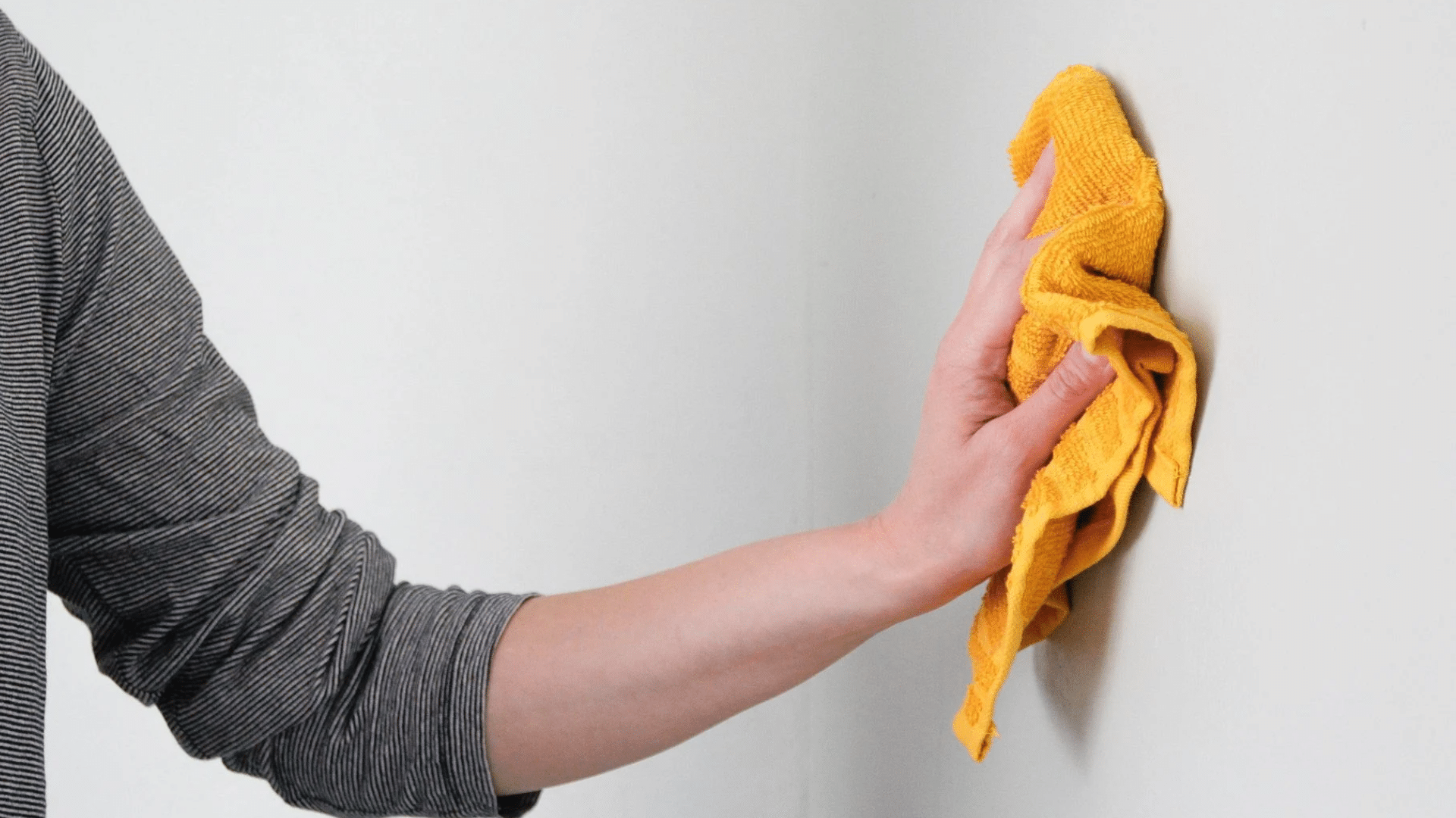 A person wearing a striped shirt uses a bright yellow cloth to wipe a plain white wall while standing in a bright room