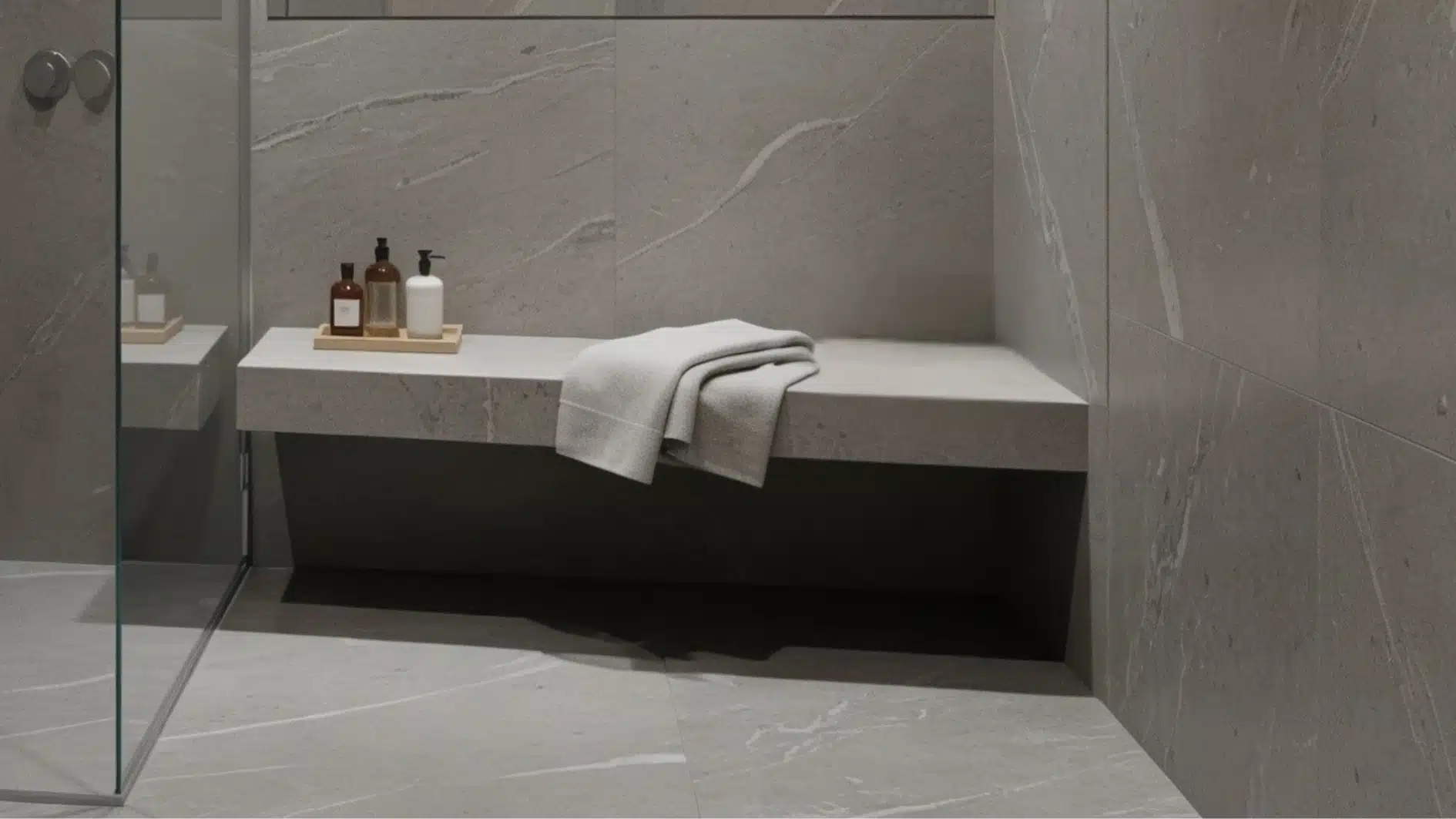 Modern walk-in shower with floor-to-ceiling grey marble-look tiles, a glass partition, a built-in stone bench with towels, and recessed ceiling lighting.