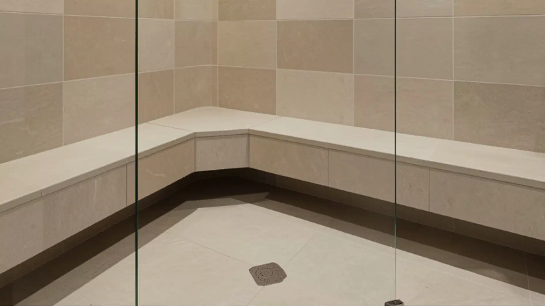 Corner built-in shower bench with light beige stone tiling and a visible glass shower partition.