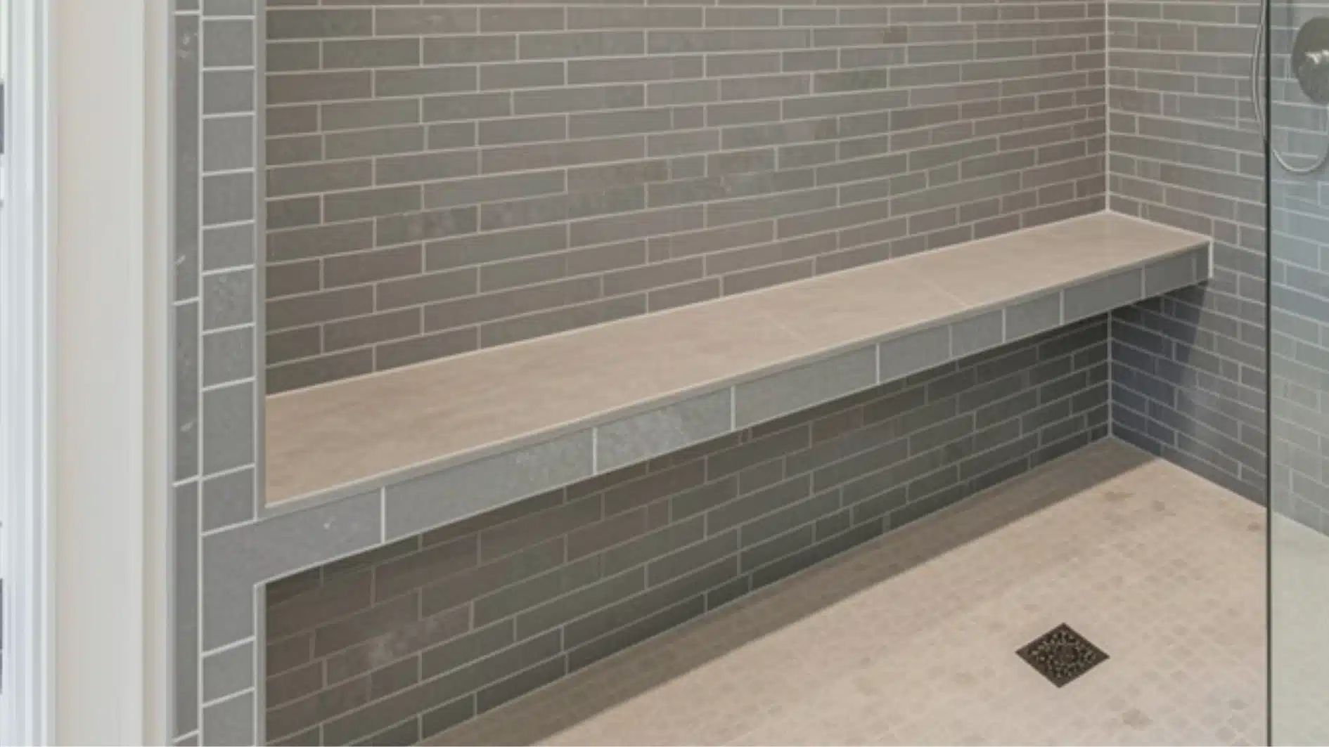 A modern shower interior featuring gray subway tile walls, a built-in light beige bench, and small square mosaic floor tiles with a dark drain.
