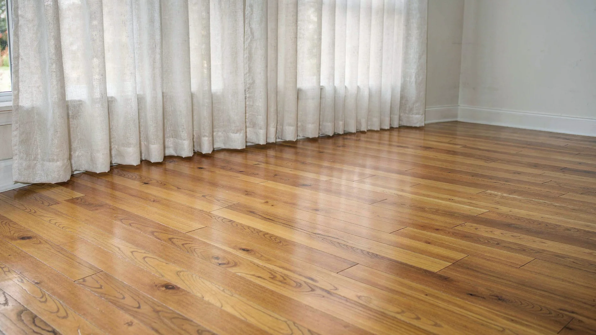 A warm honey hardwood floor extends towards sheer white curtains in a well lit room